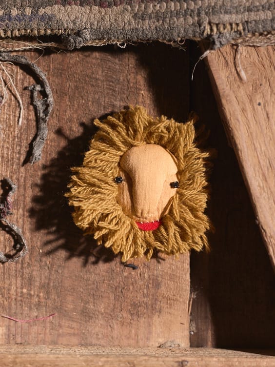 Handcrafted wooden lion face with a yarn mane mounted on a rustic wooden surface.