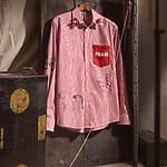 Red and white striped shirt with embroidery details and a patch reading "Fann" hanging on a rustic wooden door.