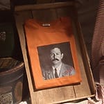 Orange t-shirt with black and white portrait of a man, displayed in a wooden tray.