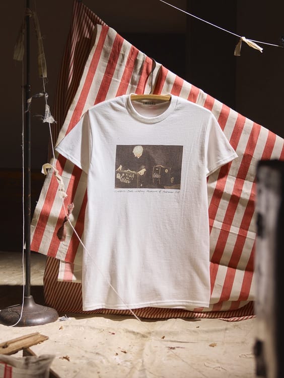 White t-shirt with abstract design displayed on hanging striped fabric backdrop indoors.