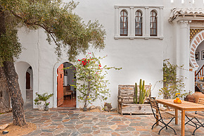 Mediterranean-style courtyard with a white wall, wooden table, chairs, cactus in a planter, and colorful flowers.