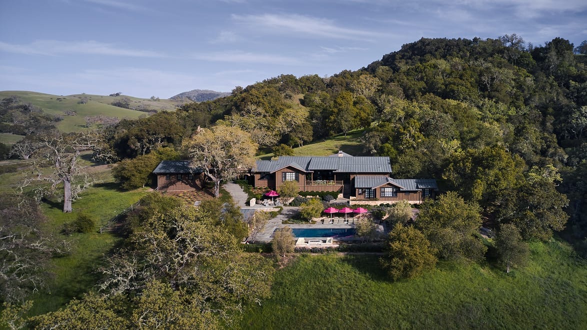 Aerial view of a large house nestled on a hillside surrounded by trees and greenery with a pool and sun umbrellas.