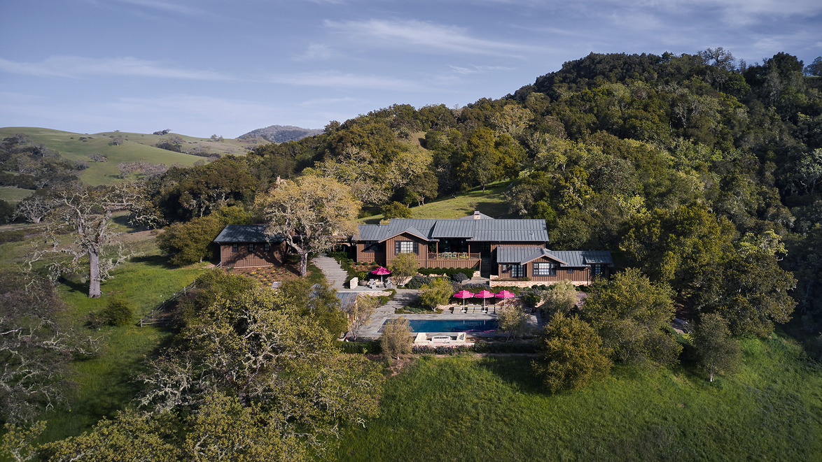 Aerial view of a large house nestled on a hillside surrounded by trees and greenery with a pool and sun umbrellas.
