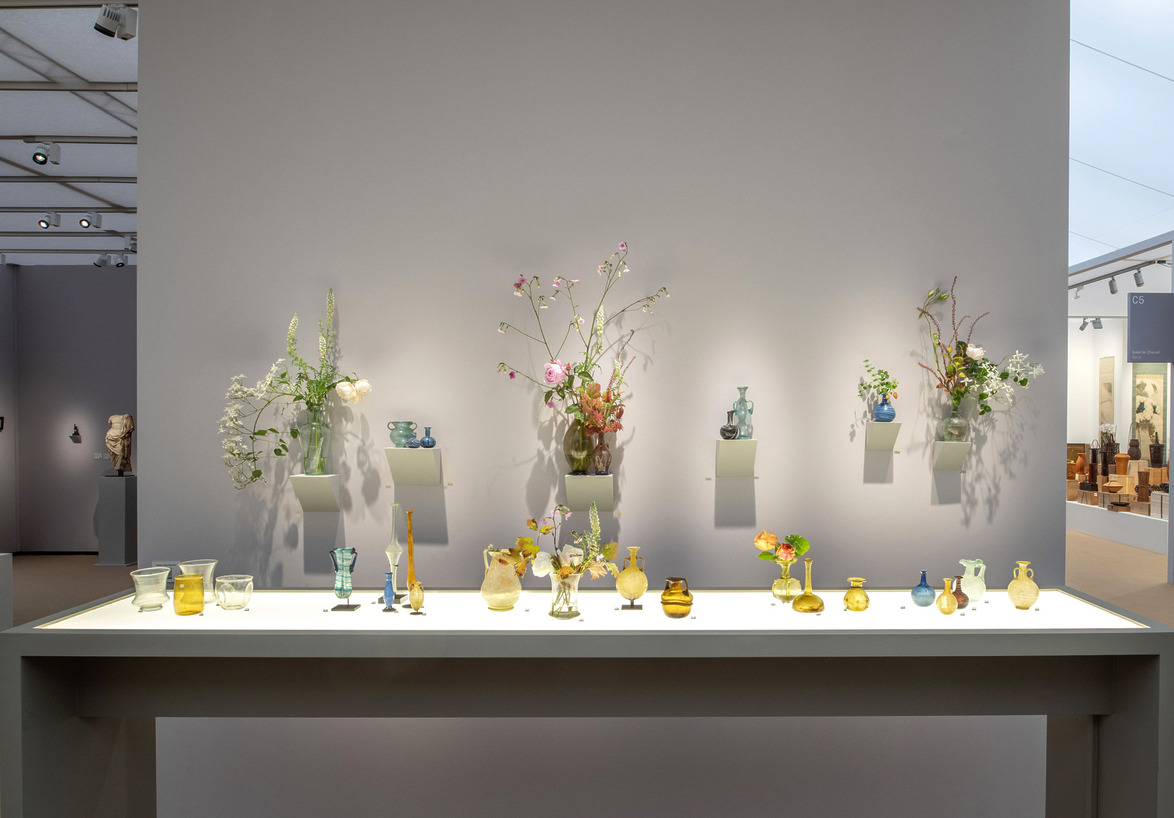 Art exhibition display with various colorful glass vases and flowers on a minimalist white table against a neutral backdrop.