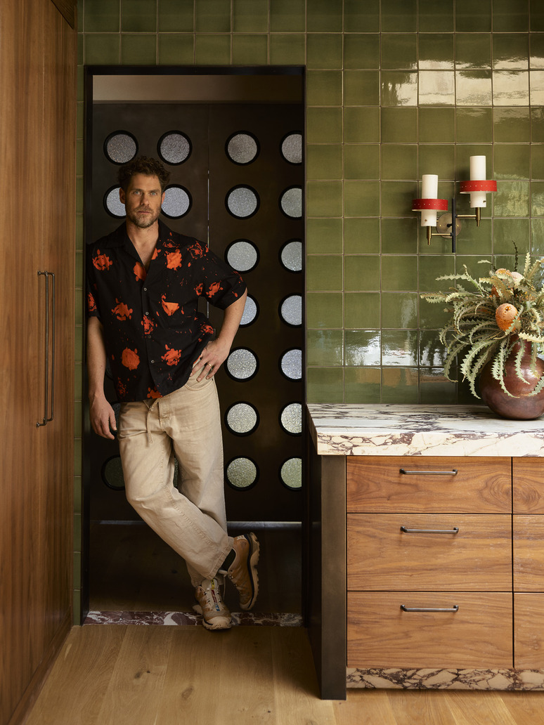 Man in floral shirt leans against a doorway in a stylish room with green tiles and wooden cabinets.