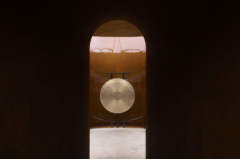 A large gong illuminated by soft light seen through an arched doorway, surrounded by a dark, minimalistic setting.