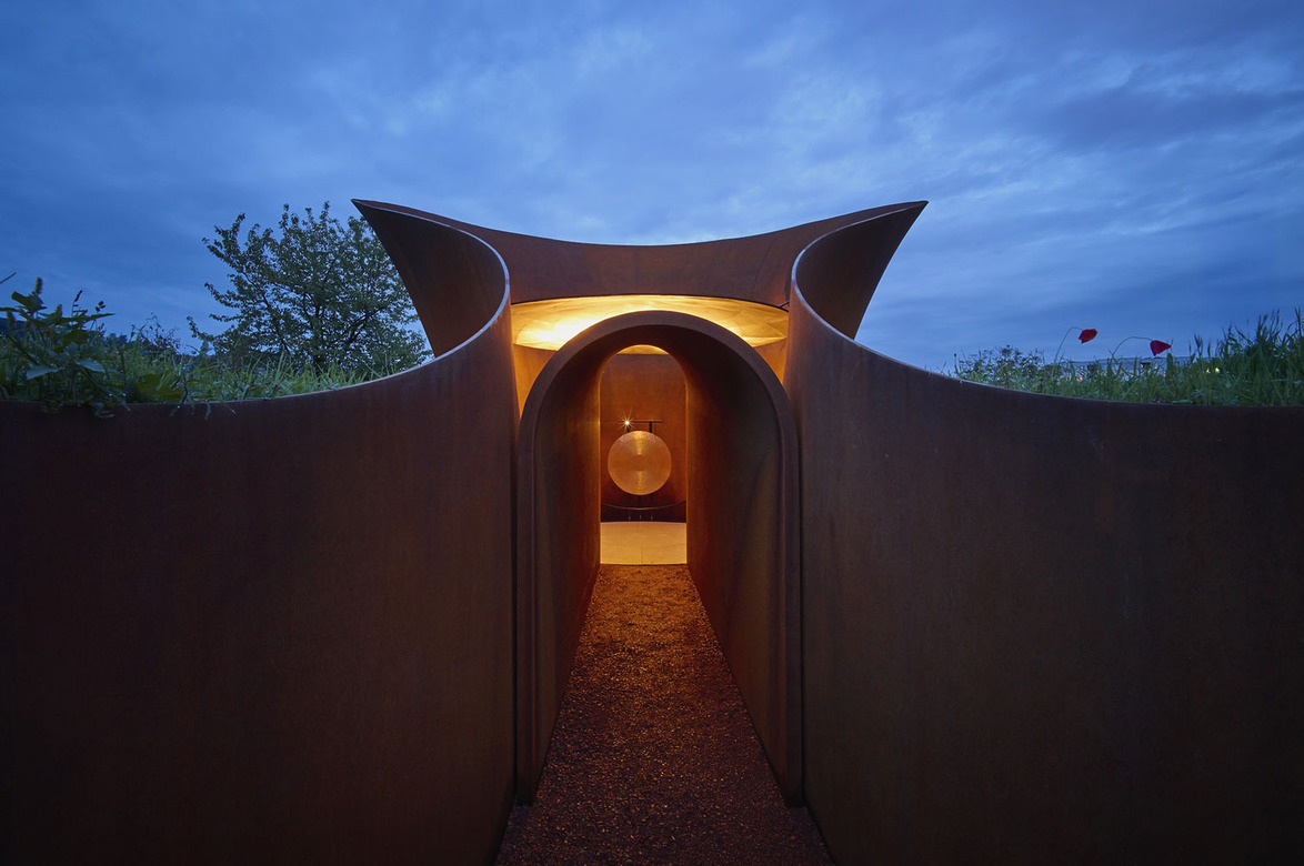 Modern outdoor art installation with curved walls and a glowing entrance during twilight hours.