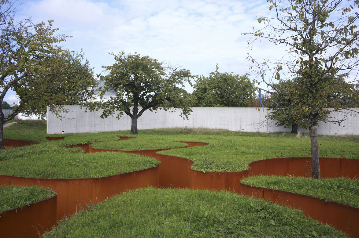 Sculpted landscape with winding grass paths and trees next to a white wall under a partly cloudy sky.