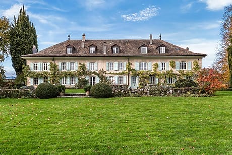 Large, elegant countryside mansion with ivy-covered walls, surrounded by lush green lawn and colorful autumn trees.