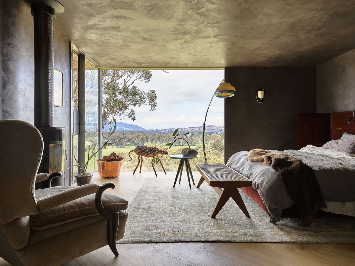 Modern bedroom with panoramic window, armchair, bench, and cozy decor overlooking a scenic landscape with mountains.