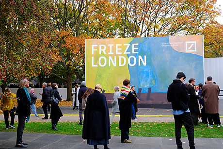 People walking around colorful Frieze London art fair sign with autumn trees in the background.