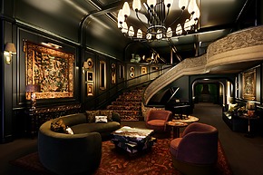 Luxurious living room with elegant staircase, chandelier, dark walls, piano, and ornate decorations.