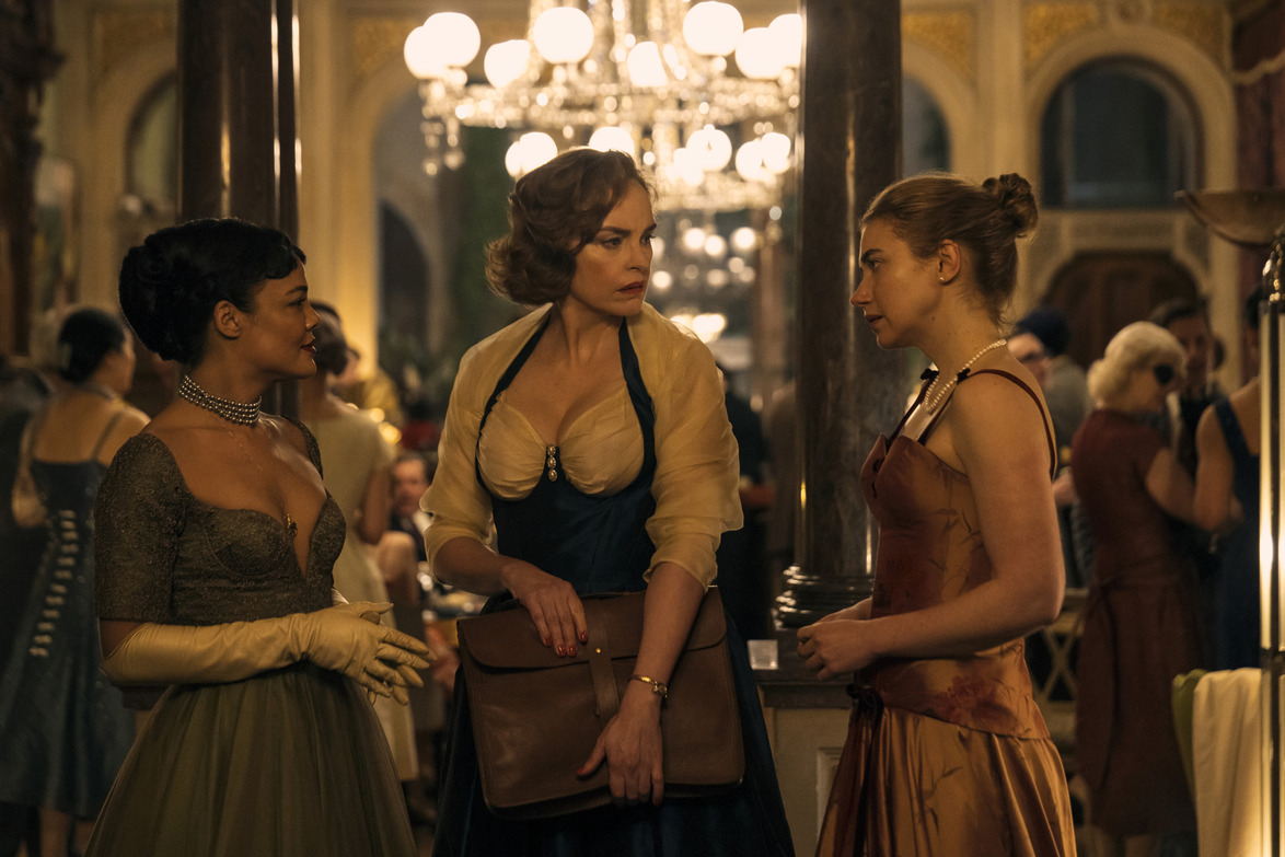 Three people in elegant attire having a conversation at a formal event with a chandelier in the background.
