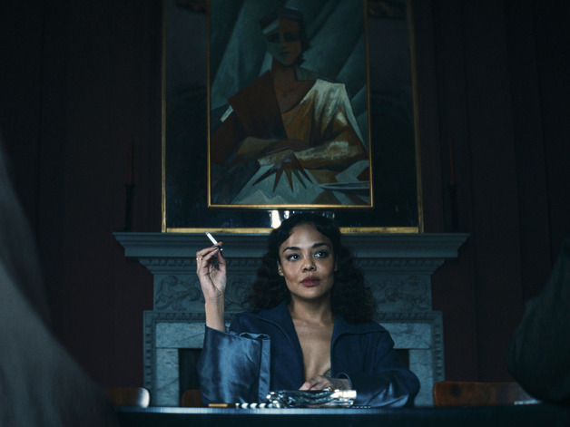 Woman in blue dress sitting at a table with a cigarette in hand, abstract portrait painting in the background.