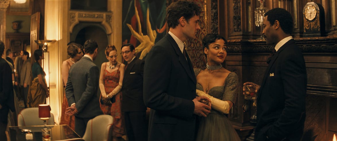 People in elegant attire converse at a sophisticated party with dim lighting and classic decor.