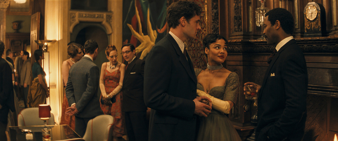 People in elegant attire converse at a sophisticated party with dim lighting and classic decor.