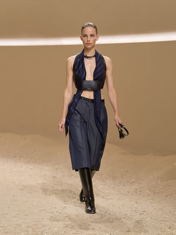 Model walking down a sandy runway in a stylish outfit with a sleeveless top and knee-length pants, holding a small accessory.
