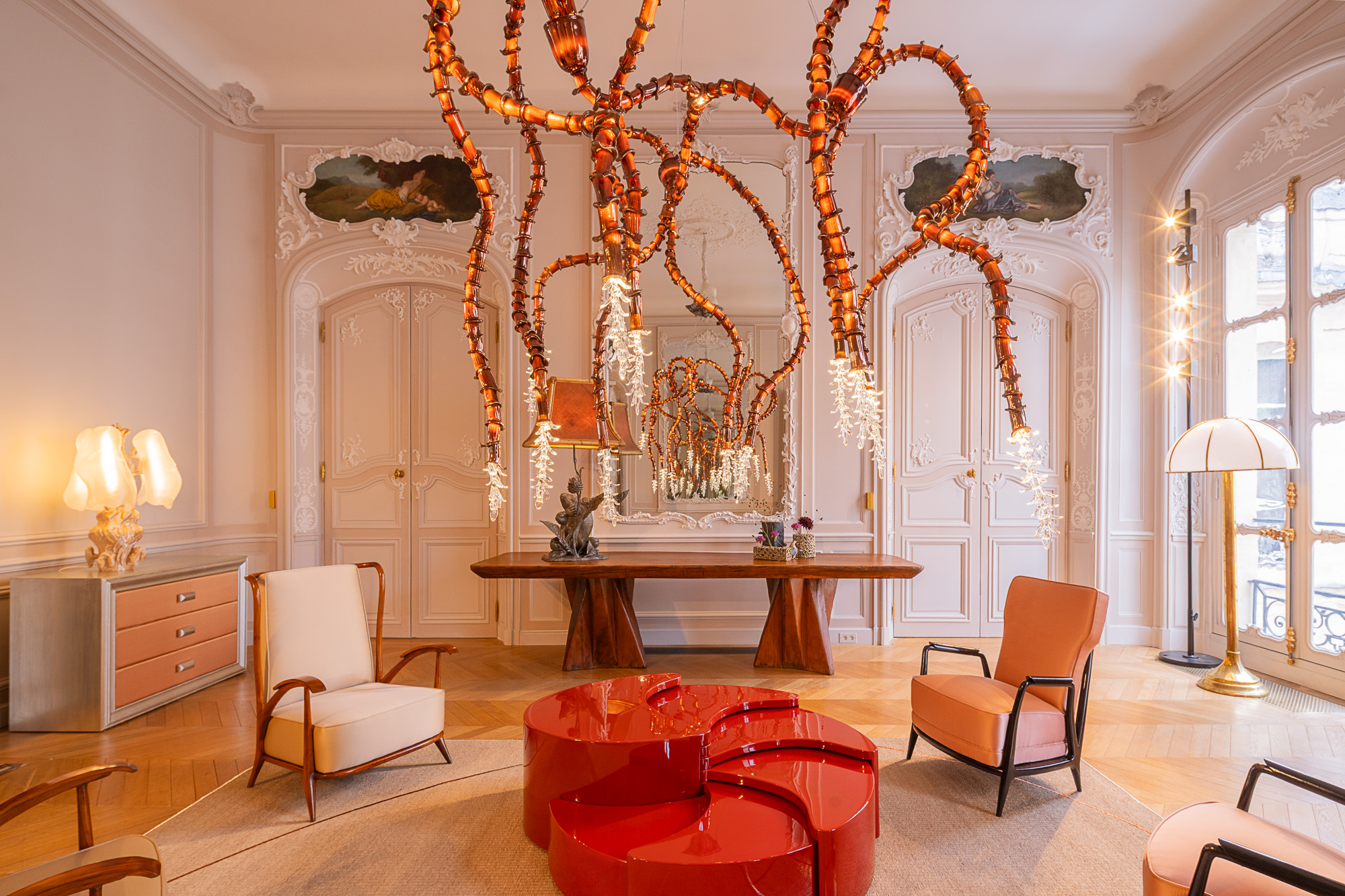 Elegant living room with ornate white walls, unique red-orange hanging light fixture, modern red tables, and stylish chairs.
