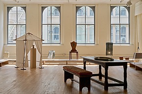 Spacious room with tall windows, wooden floors, table, bench, chair, and a small white tent structure.