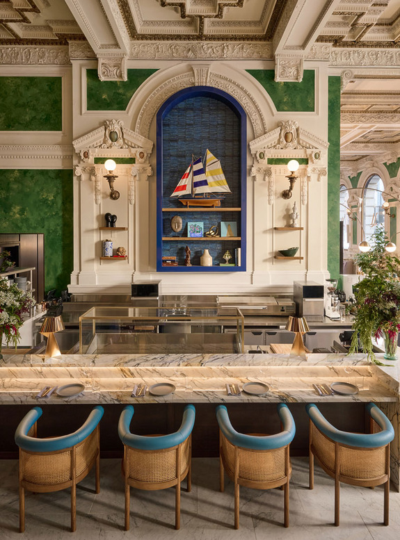 Elegant restaurant interior with marble counter, blue chairs, ornate ceiling, and nautical-themed wall art and decor.