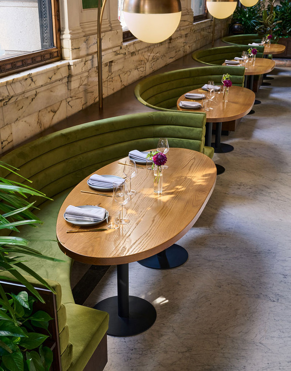 Cozy restaurant setting with green velvet booths, wooden tables set with napkins, glasses, and small flower arrangements.
