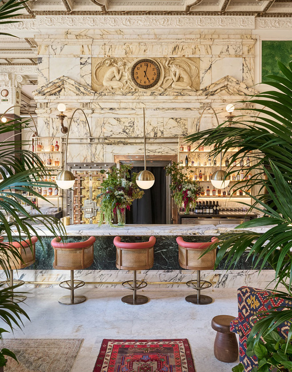 Elegant bar with marble decor, hanging lights, and plush stools surrounded by lush plants and a vintage clock on the wall.