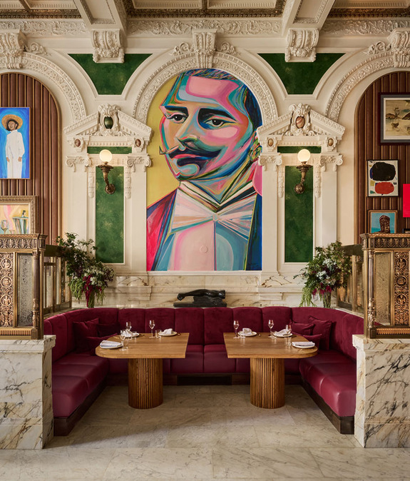 Elegant restaurant interior with ornate architecture, large colorful portrait, red velvet seating, and set dining tables.