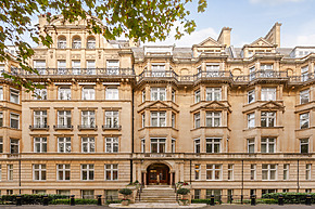Historic stone building with ornate balconies, large windows, and arched entrance surrounded by trees on a clear day.