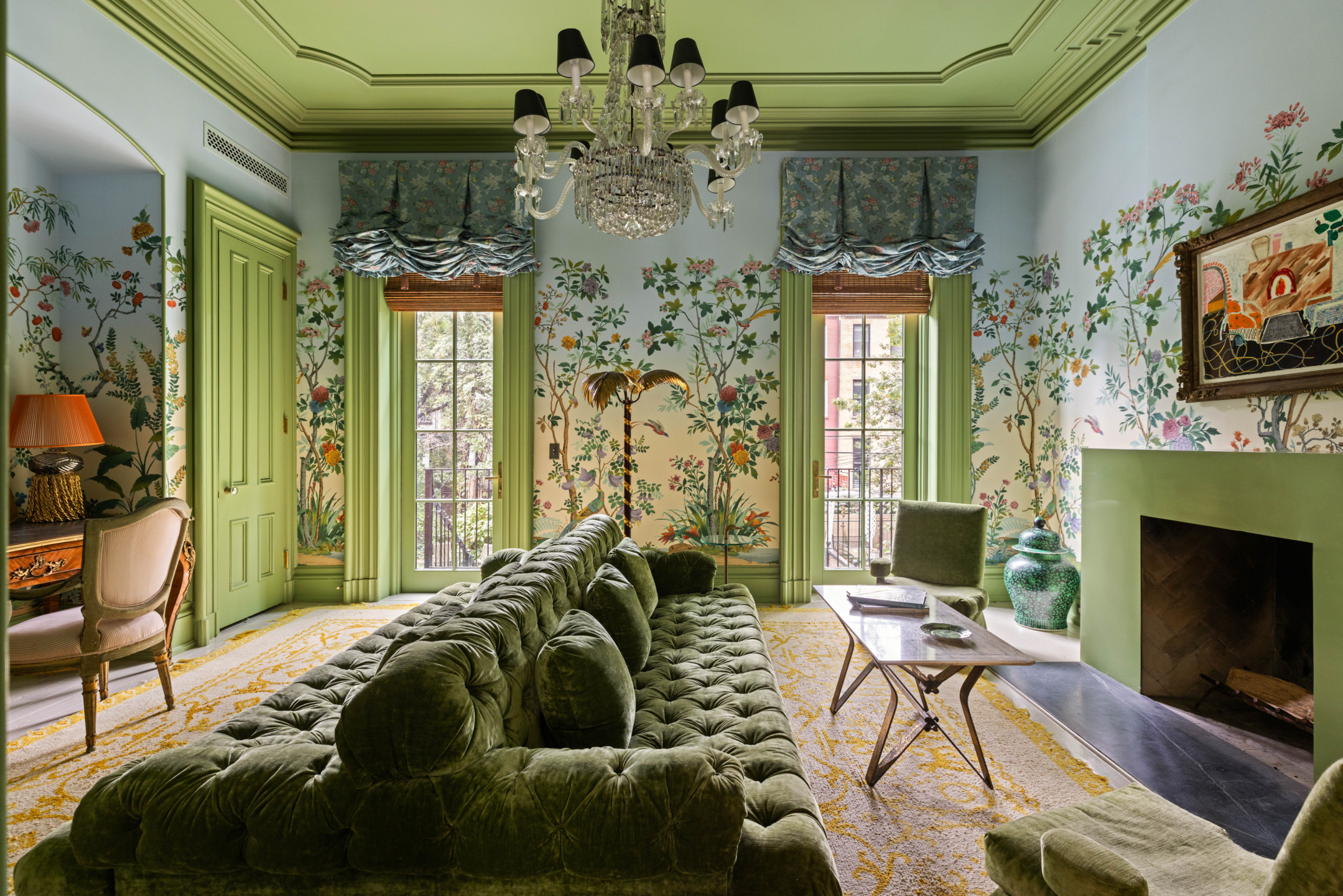Elegant living room with green velvet sofa, floral wallpaper, chandelier, fireplace, and large windows with green drapes.
