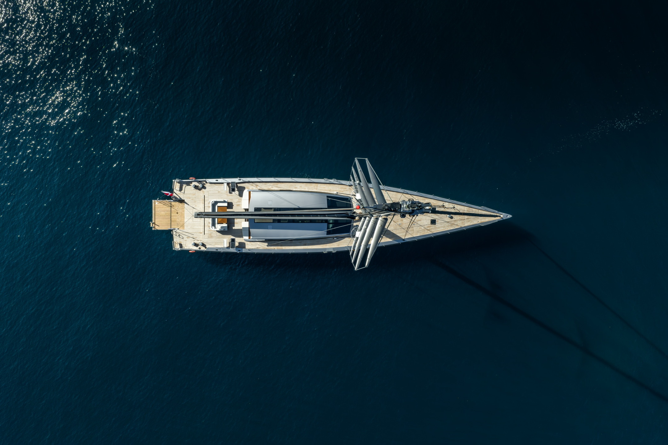 Aerial view of a sailboat floating on calm, deep blue water with sunlight reflecting off the water's surface.