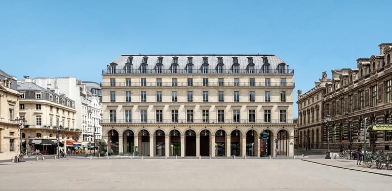 Wide view of a classic European building with arches and multiple stories, set against a clear blue sky and open courtyard.