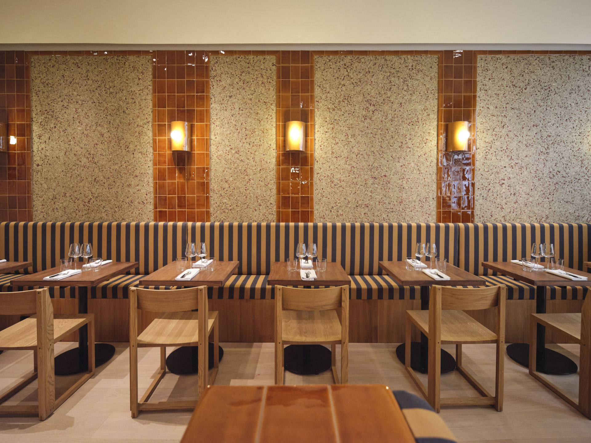 Modern restaurant interior with striped chairs, wooden tables, and soft lighting against a tiled wall backdrop.