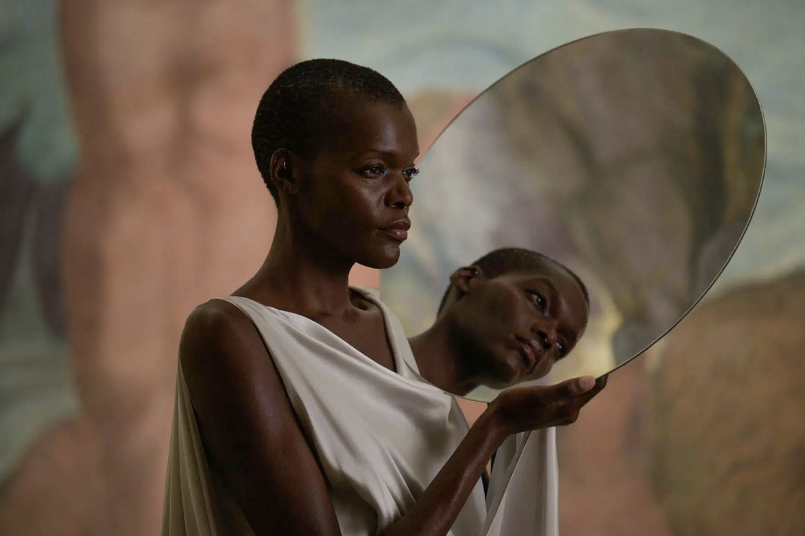 Person in white garment holding a reflective disc, creating a mirrored effect against a blurred colorful background.