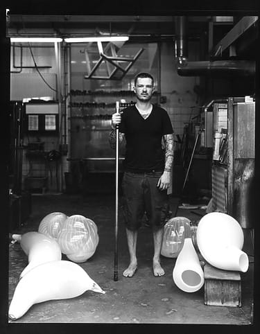 Man standing barefoot in workshop surrounded by large glass sculptures and holding a long pole.