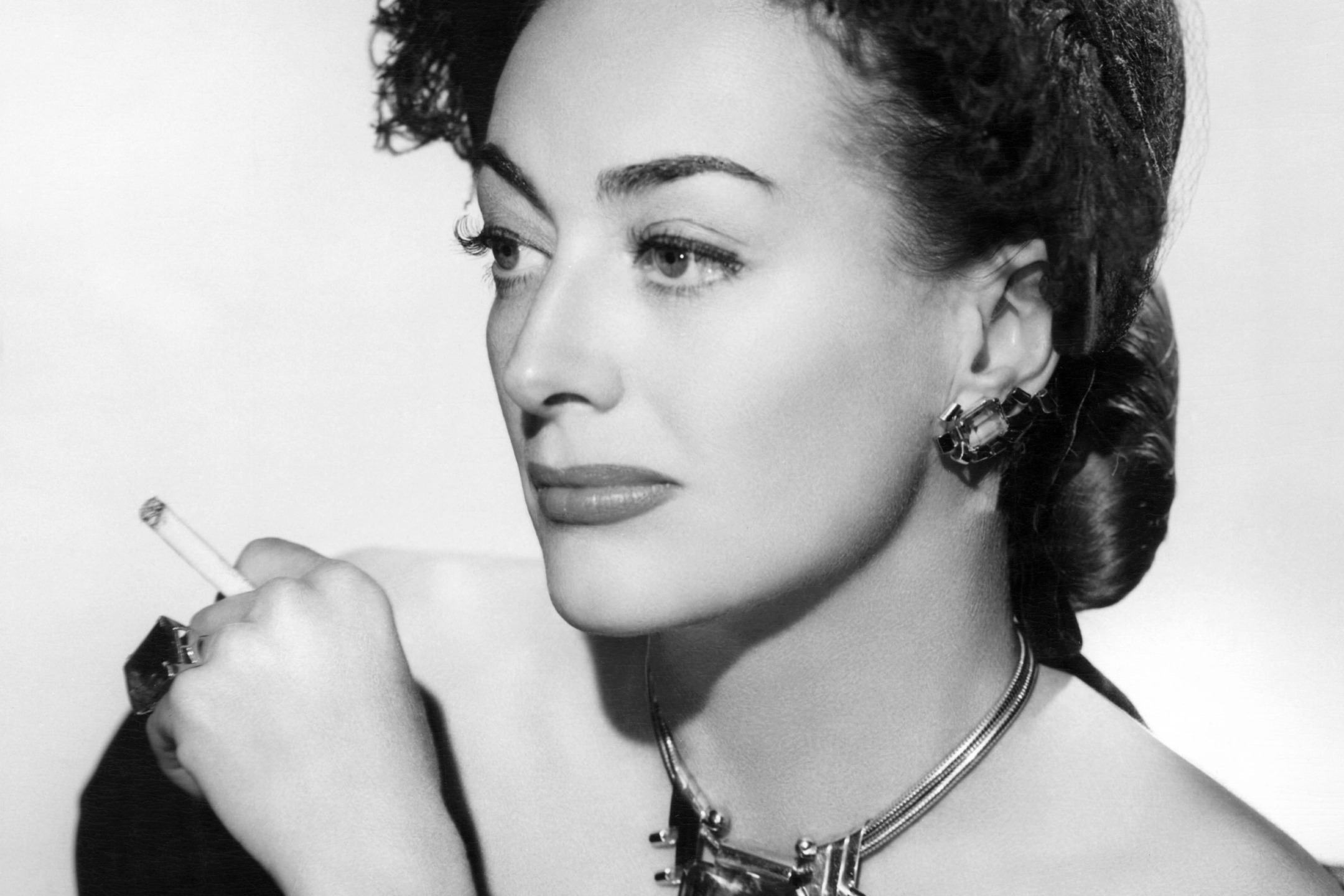 Person in black and white photo with vintage hairstyle, holding a cigarette, wearing a necklace and earrings, looking sideways.