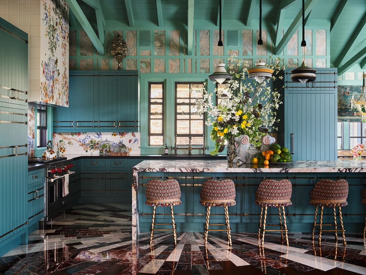Eclectic kitchen with teal cabinets, patterned bar stools, and a marble island decorated with vibrant flowers and fruits.