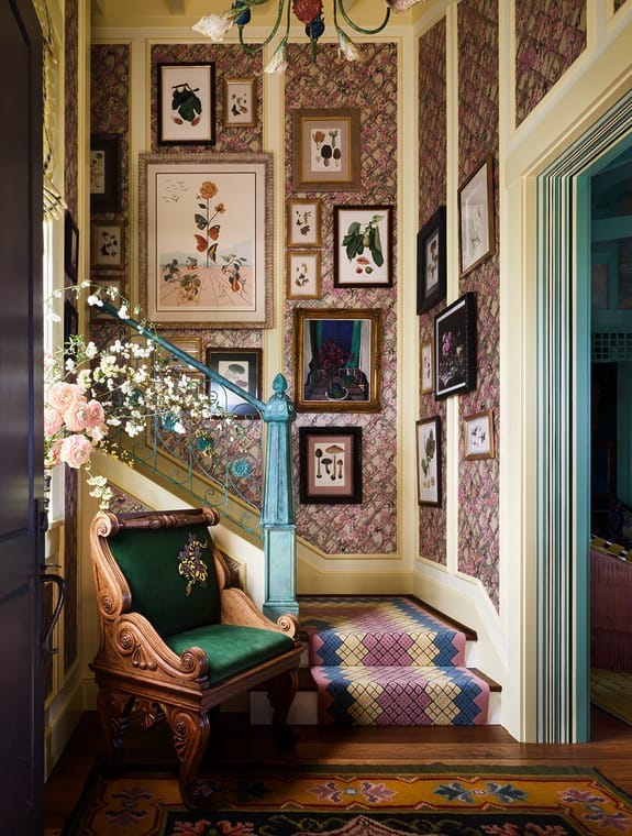 Eclectic staircase with framed art, ornate wooden chair, and patterned wallpaper in a vibrant, colorful interior.