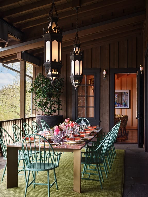 Stylish outdoor dining area with a long table set with flowers and plates, surrounded by green chairs and hanging lanterns.