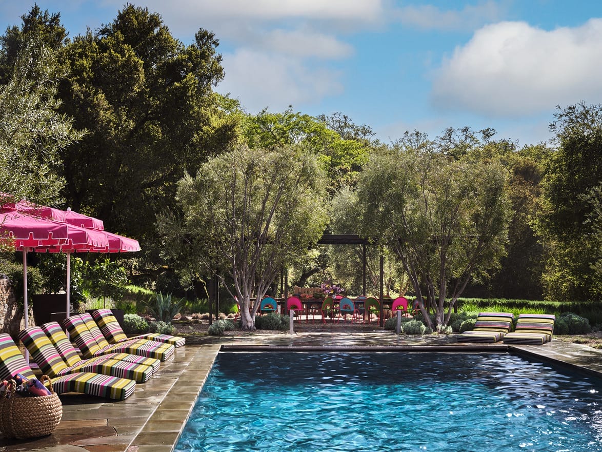 Outdoor pool surrounded by trees and striped lounge chairs under pink umbrellas on a sunny day.