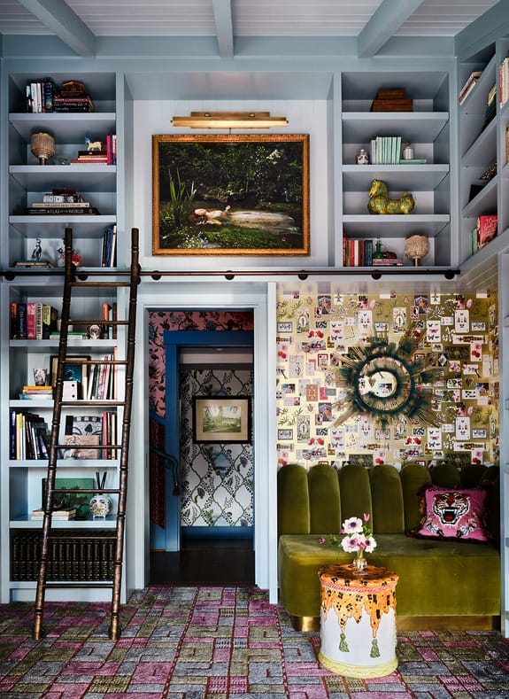 eclectic living room with green velvet sofa, bookshelves, artwork, ladder, floral wallpaper, and colorful decor items