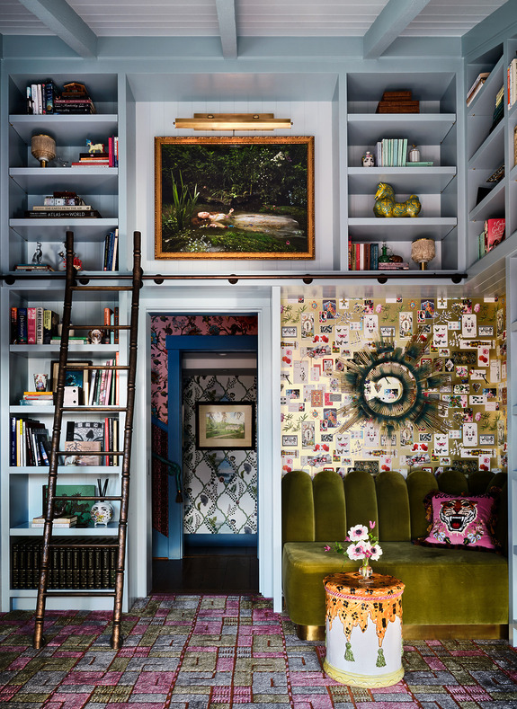 eclectic living room with green velvet sofa, bookshelves, artwork, ladder, floral wallpaper, and colorful decor items
