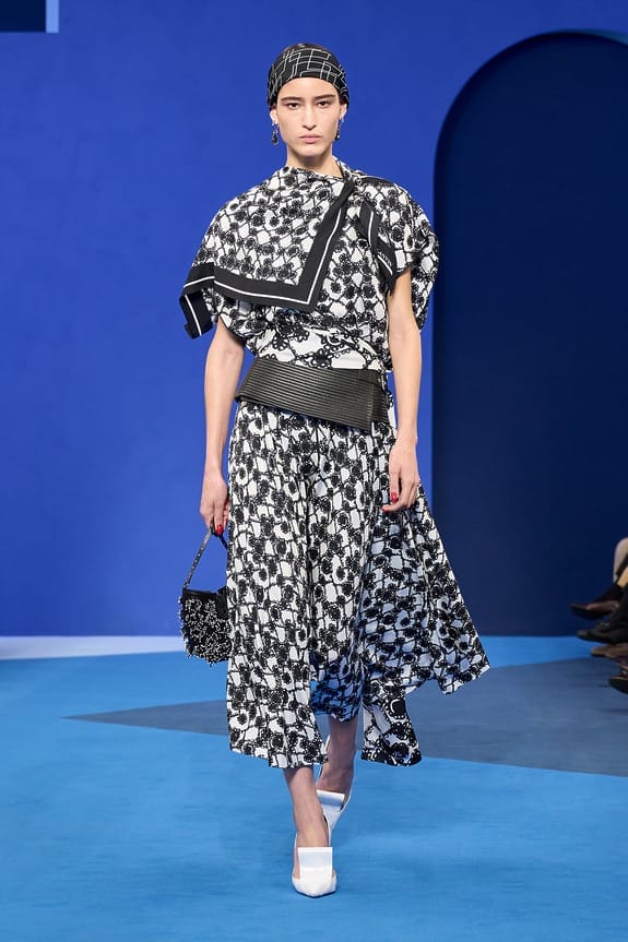 Model in a black and white patterned dress on a blue runway wearing white heels and carrying a small handbag.