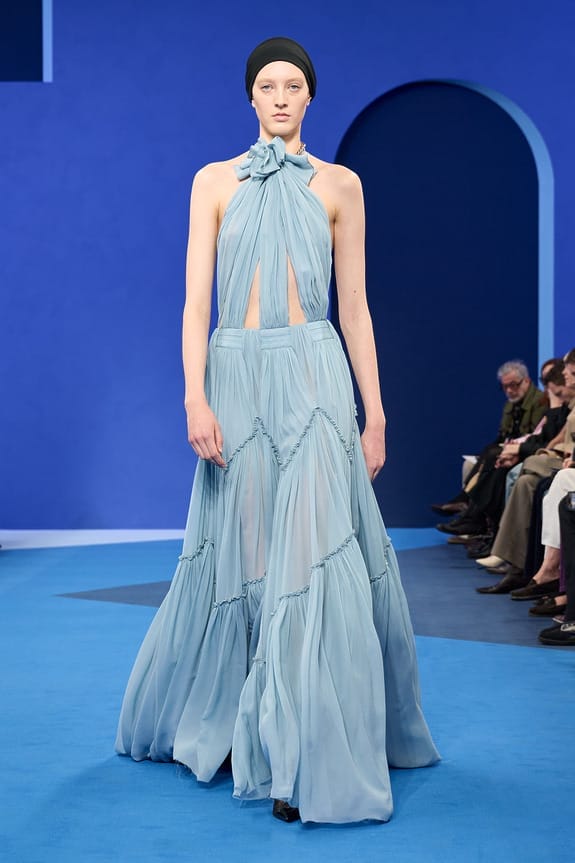 Model wearing a flowing blue dress on a runway with a blue background and audience in the background.