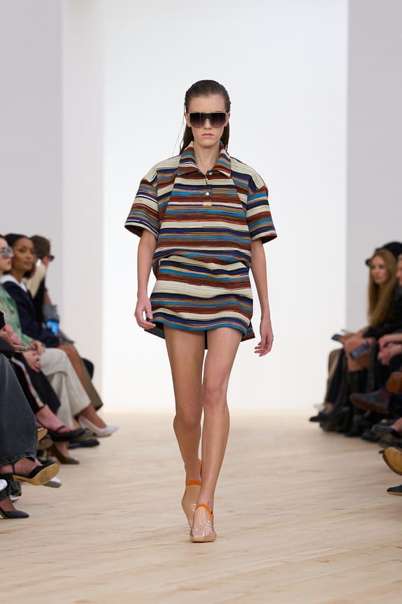 Model walking on a runway wearing a colorful striped outfit and sunglasses, with an audience seated on either side.