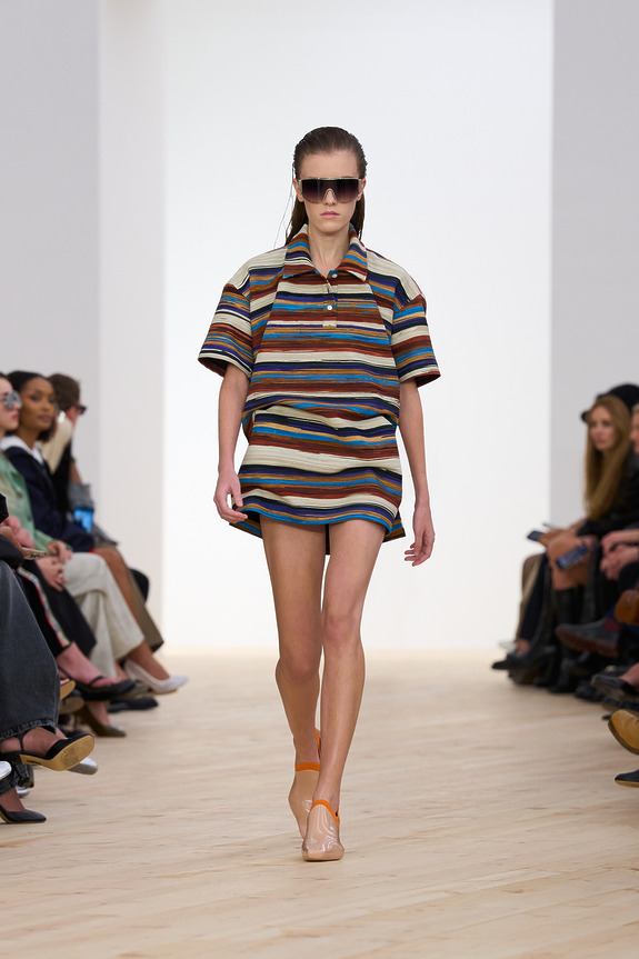Model walking on a runway wearing a colorful striped outfit and sunglasses, with an audience seated on either side.