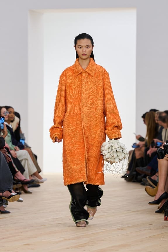 Model walking runway in bright orange textured coat holding unique handbag, surrounded by seated audience.