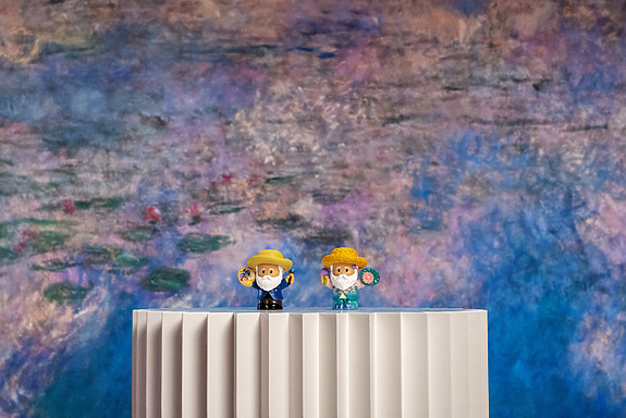 Two small figurines in front of a colorful, impressionistic backdrop on a white corrugated surface.