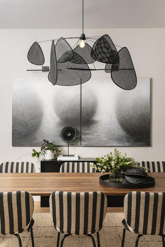 Modern dining room with abstract chandelier, striped chairs, wood table, monochrome artwork, and decorative plants.