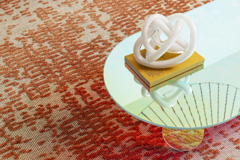 Glass coffee table with a knot sculpture on top, colorful patterned rug underneath, casting reflections on the table surface.