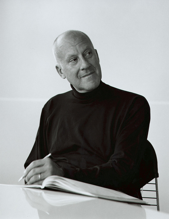 Man in black turtleneck sitting at a desk with an open notebook, looking to the side with a thoughtful expression.